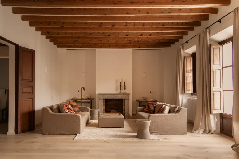 Salón con dos sofás, chimenea de obra y vigas de madera originales en el proyecto 1906 de Sa Dutxeta en Sóller, Mallorca